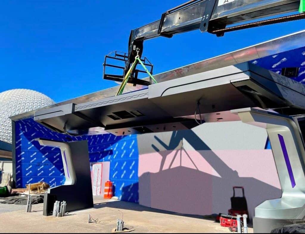 A large metal structure is lifted into place by crane at EPCOT, right in front of the vibrant purple wall of the upcoming Guardians of the Galaxy: Cosmic Rewind attraction, with Spaceship Earth's dome and a bright blue sky completing this exciting construction scene.