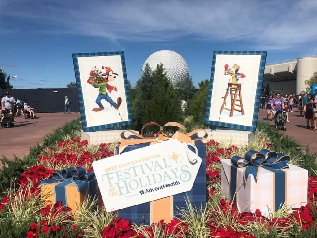 Colorful gift boxes and a festive sign welcome guests to EPCOT’s International Festival of the Holidays, featuring cheerful Goofy holiday artwork and vibrant floral displays.