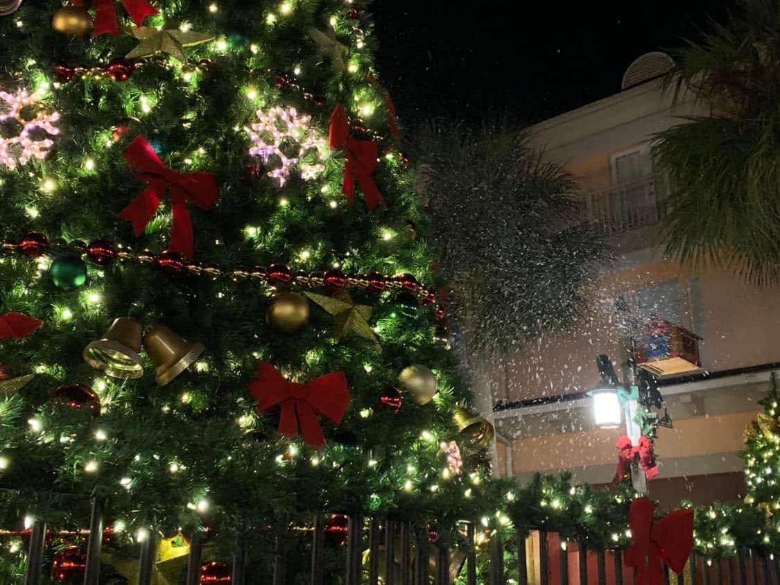 A towering Christmas tree adorned with twinkling lights, colorful ornaments, and bright red bows stands in front of a themed park building at night, setting the perfect scene for holiday festivities at the heart of the resort.
