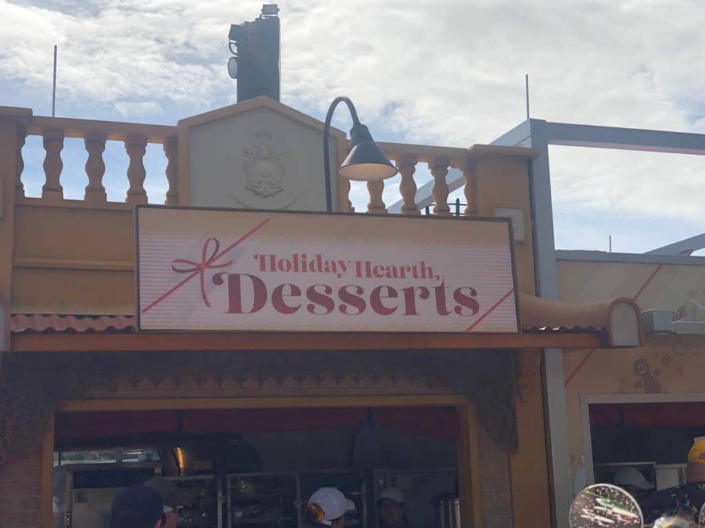 Holiday Hearth Desserts stand at EPCOT International Festival of the Holidays, featuring its festive sign, a glowing lamp overhead, guests gathered below enjoying the seasonal treats, and clear blue sky completing the magical atmosphere of Disney’s beloved Holiday Kitchens.