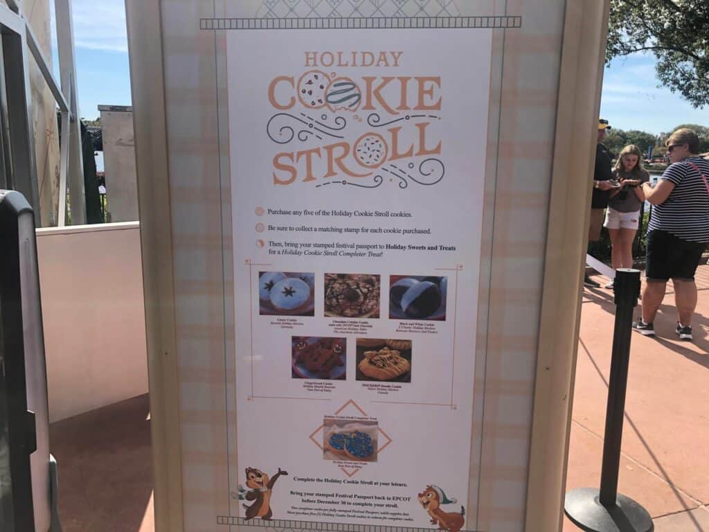 Holiday Cookie Stroll sign at EPCOT International Festival of the Holidays, showcasing step-by-step participation details and colorful cookie graphics; guests savoring festive treats and exploring World Showcase in the background.