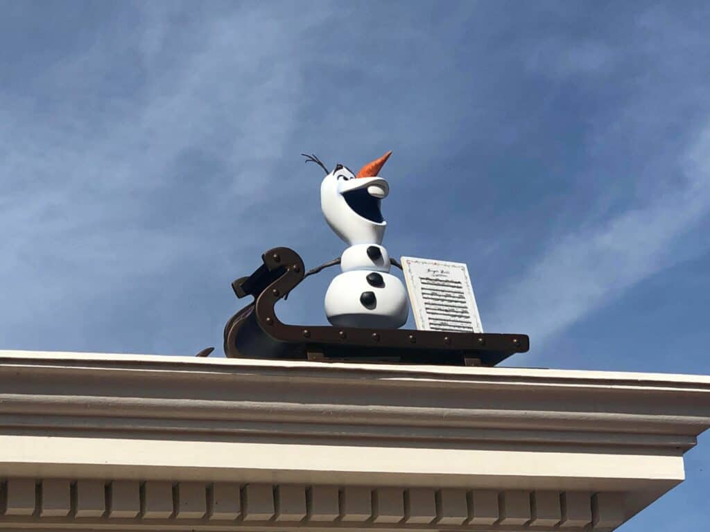 A cheerful snowman with a classic carrot nose lounges on a sleigh atop an EPCOT building, set against a bright blue sky, adding whimsical holiday magic to the park’s festive atmosphere.