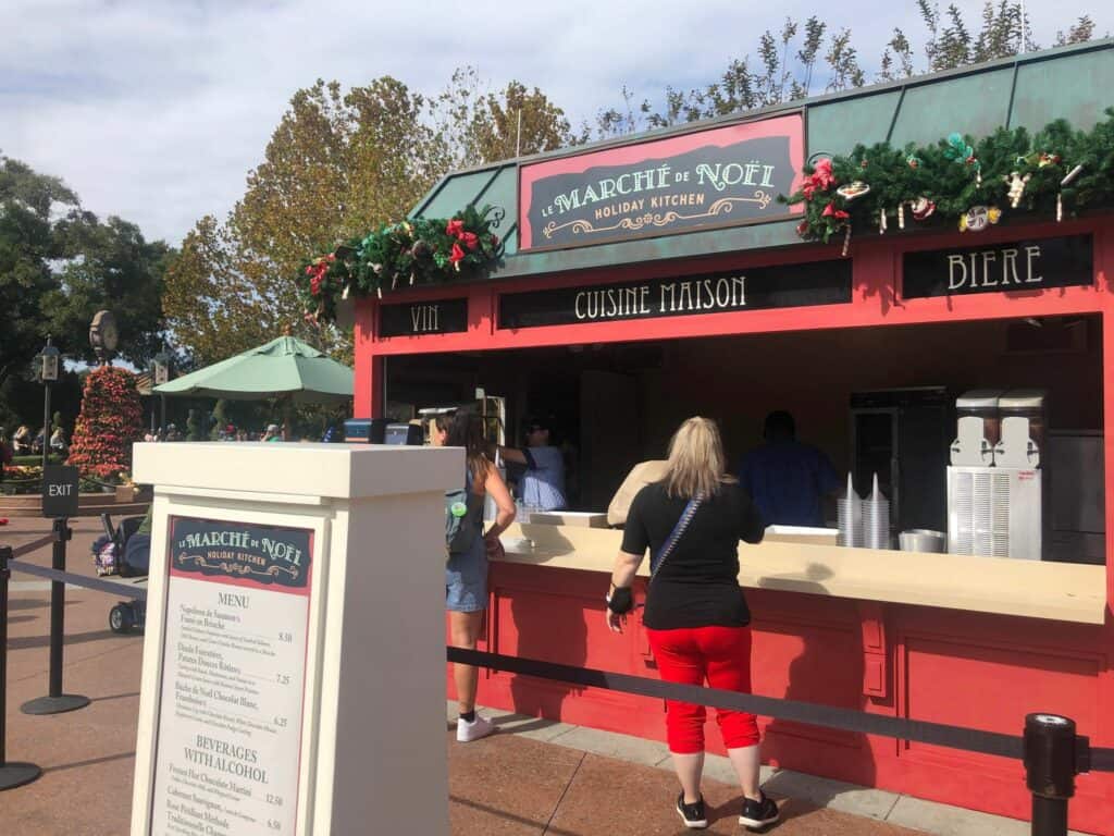 Guests line up to sample French-inspired holiday treats at a festively decorated EPCOT Holiday Kitchen, adorned with garlands and classic French signage, during the EPCOT International Festival of the Holidays.