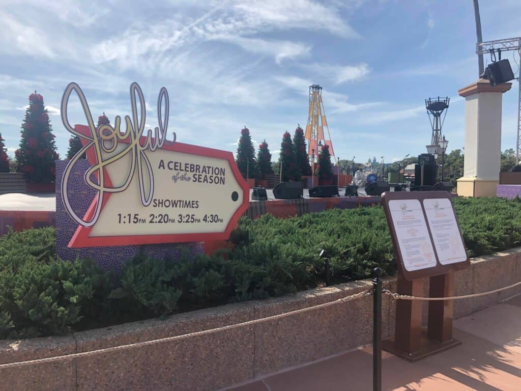 EPCOT outdoor stage featuring the "Joyful, A Celebration of the Season" sign with performance times and a menu display, spotlighting live holiday entertainment at the International Festival of the Holidays.