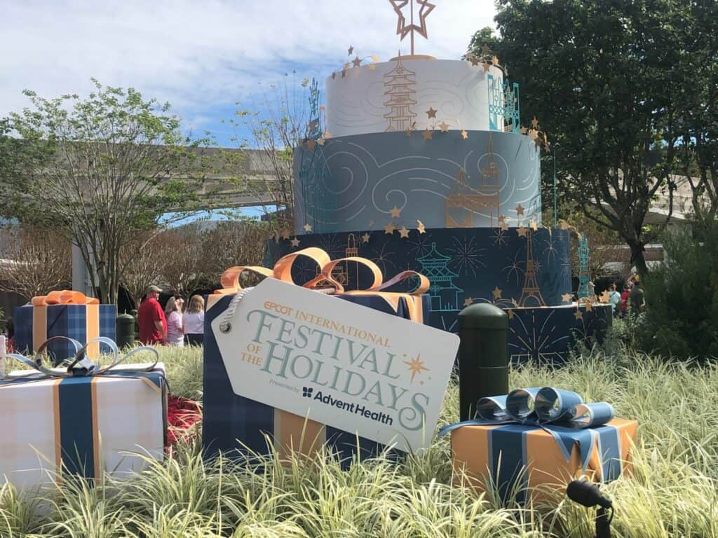 A festive holiday cake and colorful presents sit beneath a "Festival of the Holidays" sign at EPCOT, surrounded by guests savoring delicious treats from the Holiday Kitchens and soaking up the seasonal magic.