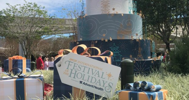 A festive holiday cake and colorful presents sit beneath a "Festival of the Holidays" sign at EPCOT, surrounded by guests savoring delicious treats from the Holiday Kitchens and soaking up the seasonal magic.