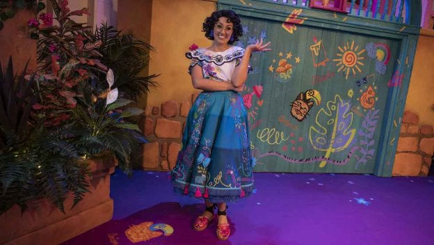 Mirabel beams in a colorful outfit, striking a playful pose amid lush plants and an enchanting, vibrant backdrop inspired by the world of Encanto at Disney California Adventure Park.