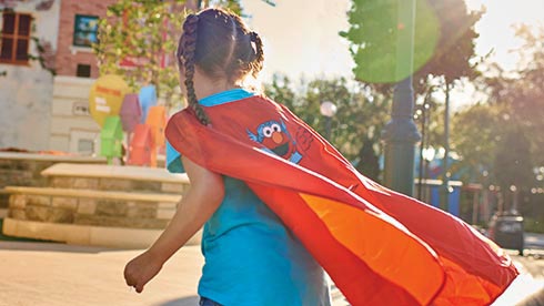 A young Sesame Street fan sporting braids steps out in a vibrant red Elmo cape, enjoying the sunshine and excitement of Kid’s Weekend—ready to explore all the Sesame Street-themed adventures the park has to offer.