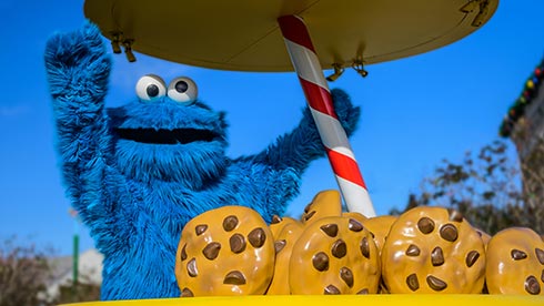Cookie Monster from Sesame Street waves enthusiastically behind giant chocolate chip cookies under a striped pole, with blue sky overhead—capturing the lively spirit of SeaWorld’s Kid’s Weekend celebration.
