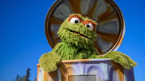 Oscar the Grouch emerges from his classic trash can in front of a vivid blue sky, creating a lively Sesame Street moment that’s sure to delight families and kids visiting the park on Kid's Weekend.