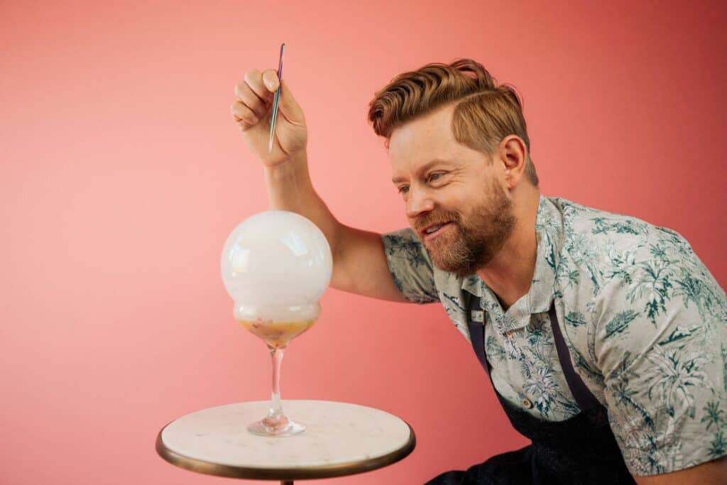 A cheerful guest at Florida Kitchen by Richard Blais in the Hyatt Regency Grand Cypress smiles as he pops a dramatic bubble topping his cocktail, sitting at a cozy round table against a whimsical pink backdrop, capturing the restaurant’s signature playful culinary flair.