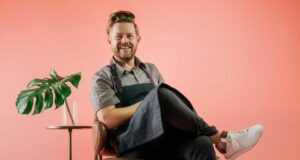 Celebrity chef Richard Blais beams with enthusiasm, lounging cross-legged in a vibrant, pink-themed setting reminiscent of Four Flamingos: A Florida Kitchen—capturing the playful energy and inviting atmosphere found in top Orlando entertainment spots. A leafy plant on the nearby table adds to the festive, tropical vibe.