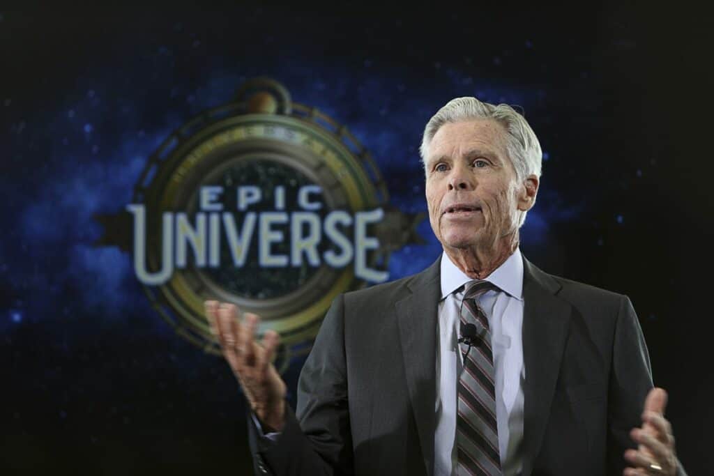 Universal CEO Tom Williams takes the stage in a suit with the "Epic Universe" logo illuminated behind him, delivering his retirement announcement during this landmark moment in Universal Orlando Resort history.