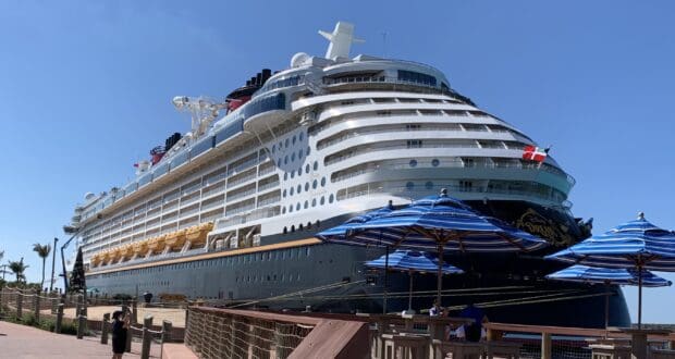 Disney Cruise Line ship docked at port beneath a clear blue sky, with iconic striped umbrellas offering shade as guests stroll by—some chatting about boarding protocols or checking email before their magical voyage begins.