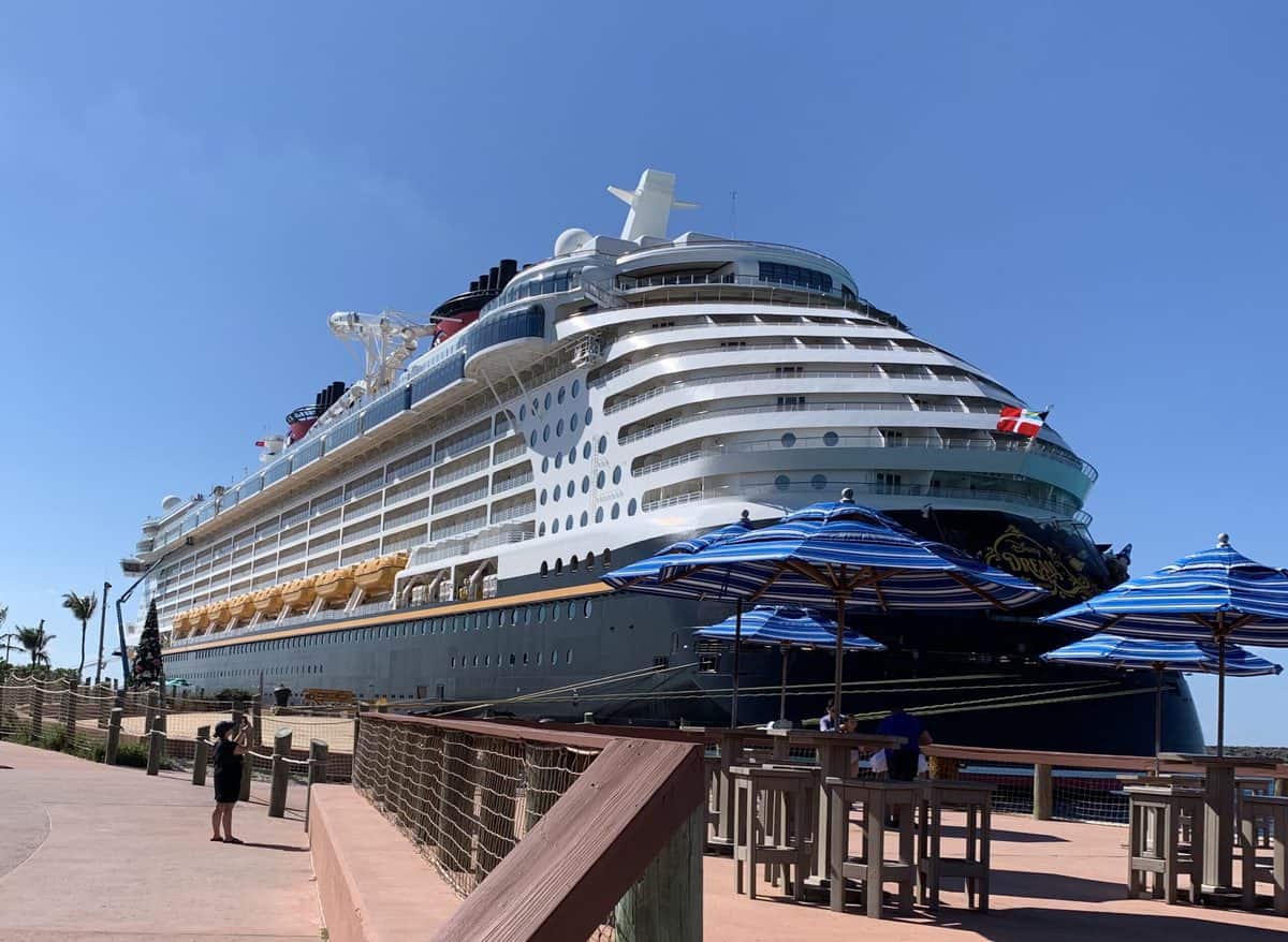 Disney Cruise Line ship docked at port beneath a clear blue sky, with iconic striped umbrellas offering shade as guests stroll by—some chatting about boarding protocols or checking email before their magical voyage begins.