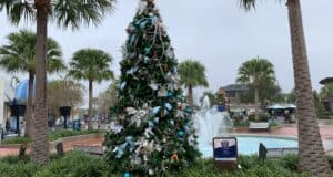 A dazzling Christmas tree sparkles between swaying palm trees beside the iconic fountain on Main Street, creating a magical holiday atmosphere perfect for seasonal dining celebrations at the park—even with clouds overhead.