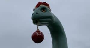 Gertie the Dinosaur at Disney’s Hollywood Studios rocks a festive Santa hat and playfully holds a red "50" ornament in her mouth beneath a cloudy sky, celebrating the park’s holiday season and 50th anniversary magic.