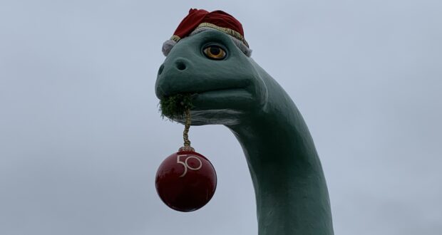 Gertie the Dinosaur at Disney’s Hollywood Studios rocks a festive Santa hat and playfully holds a red "50" ornament in her mouth beneath a cloudy sky, celebrating the park’s holiday season and 50th anniversary magic.