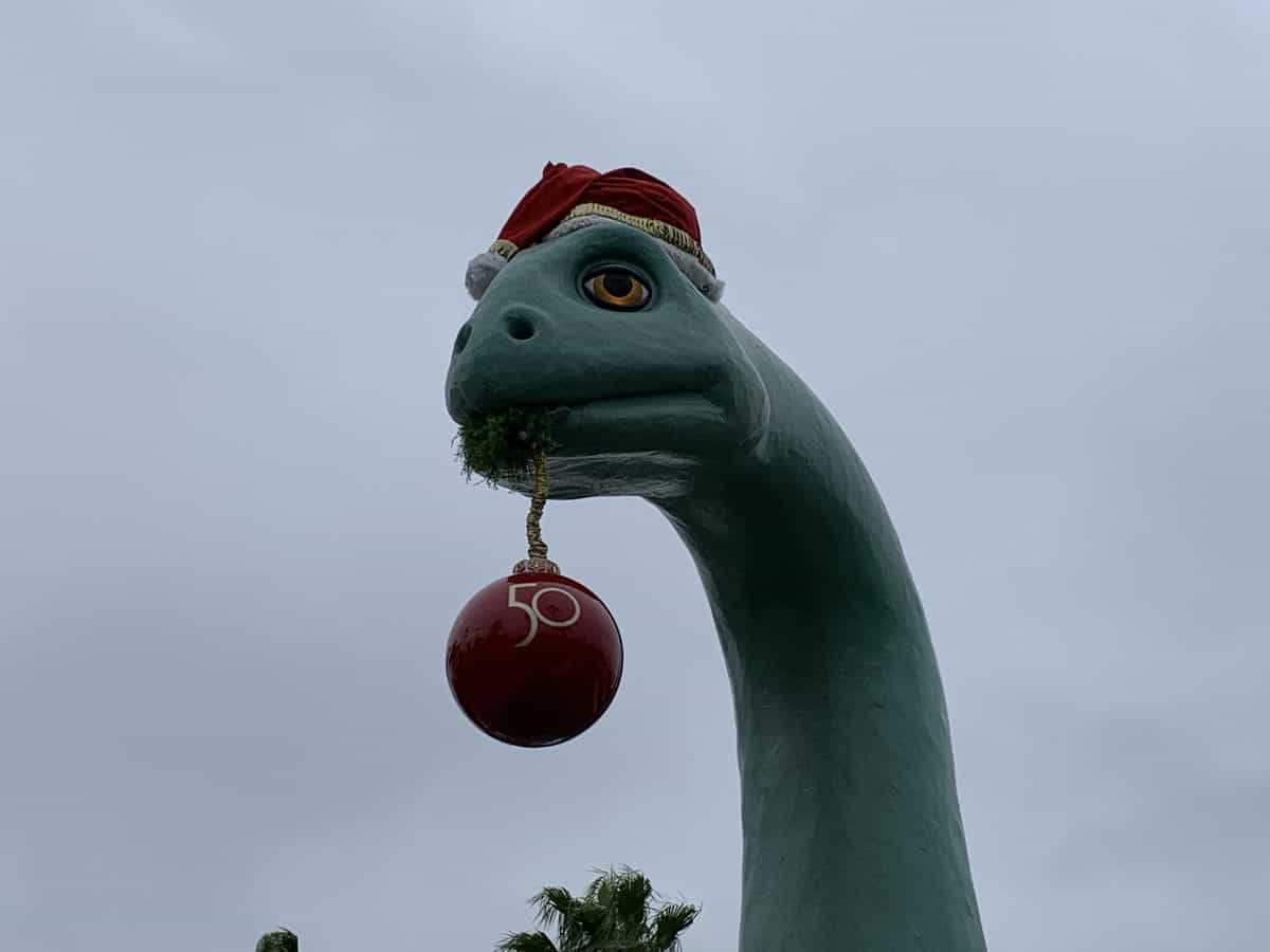 Gertie the Dinosaur at Disney’s Hollywood Studios rocks a festive Santa hat and playfully holds a red "50" ornament in her mouth beneath a cloudy sky, celebrating the park’s holiday season and 50th anniversary magic.