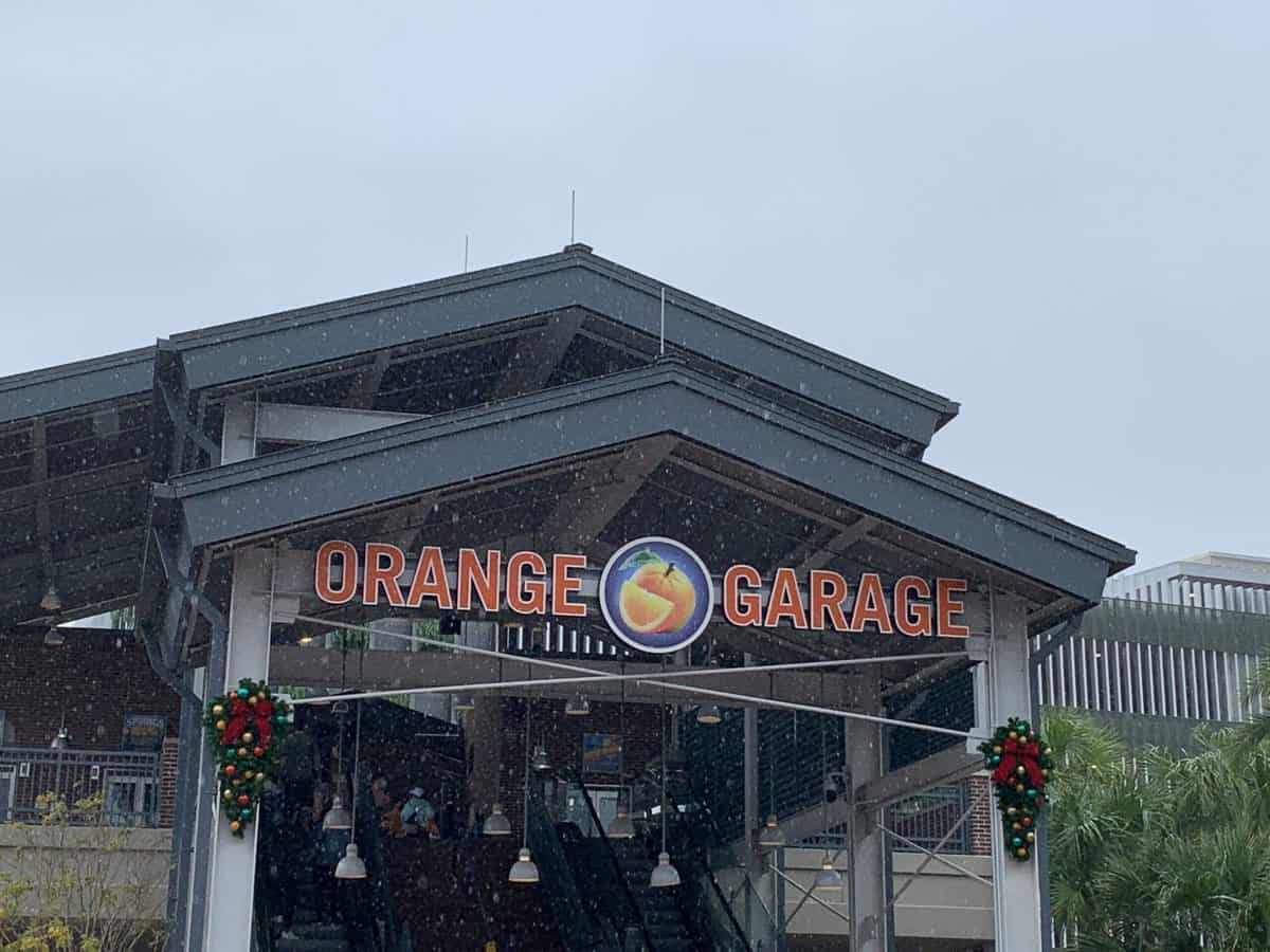 The Orange Garage entrance sign at Disney Springs, featuring its signature peach logo and festive wreath decorations, stands out beneath a cloudy sky—your go-to landmark for easy parking before diving into the shops, dining, and entertainment of Disney Springs.