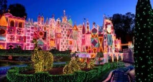 A colorful, whimsical building and topiary animals are covered in glowing holiday lights at dusk, set against a deep blue sky, creating a festive and enchanting nighttime scene.