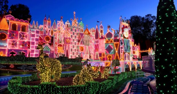 A colorful, whimsical building and topiary animals are covered in glowing holiday lights at dusk, set against a deep blue sky, creating a festive and enchanting nighttime scene.