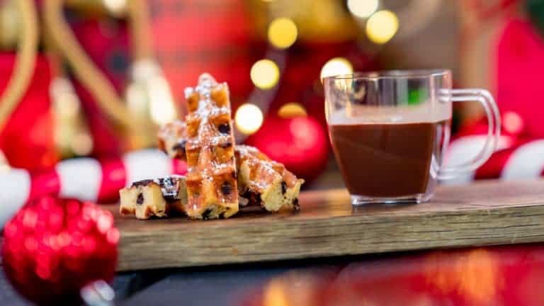 Hot chocolate in a glass cup and a stack of waffles served on a wooden board, surrounded by holiday decorations, all set against the whimsical backdrop of Disneyland’s It’s a Small World attraction.