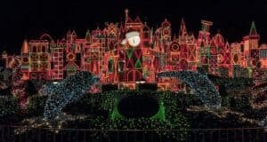 It’s a Small World’s whimsical facade sparkles with vibrant Christmas lights against the night sky, surrounded by lush greenery and playful dolphin sculptures, creating the beloved holiday magic Disney fans adore.