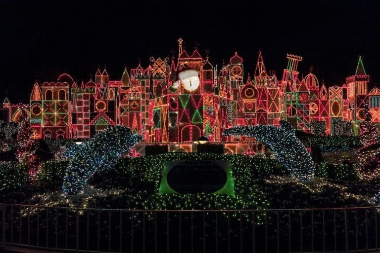 It’s a Small World’s whimsical facade sparkles with vibrant Christmas lights against the night sky, surrounded by lush greenery and playful dolphin sculptures, creating the beloved holiday magic Disney fans adore.