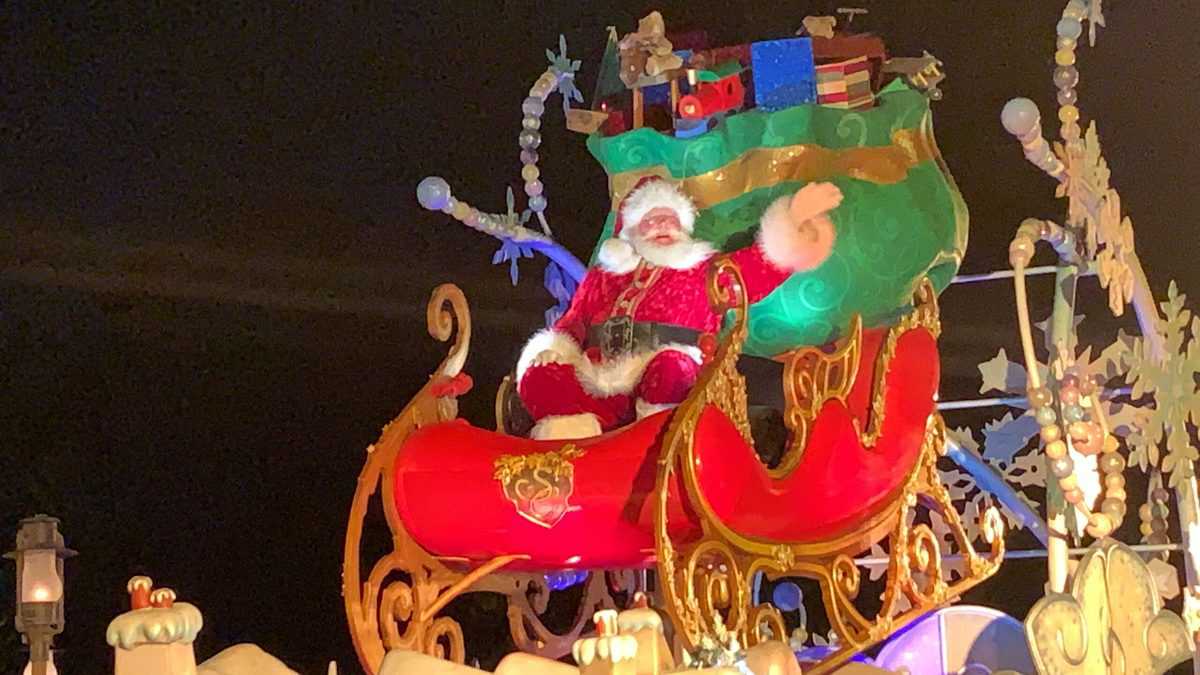 Santa Claus waves energetically from his red sleigh overflowing with gifts as he glides through a sparkling nighttime parade, surrounded by dazzling festive decorations, capturing the enchanting spirit of Mickey’s Very Merry Christmas Party at Magic Kingdom.