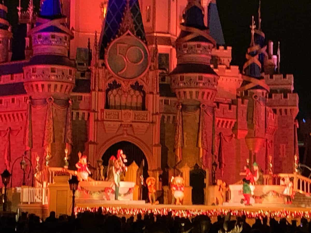 The iconic Cinderella Castle at Magic Kingdom shines with vibrant lights and festive décor, as beloved Disney characters perform in elaborate holiday costumes beneath a giant "50" anniversary sign—a scene capturing the magic of both Disney Very Merriest After Hours and Mickey’s Very Merry Christmas Party.