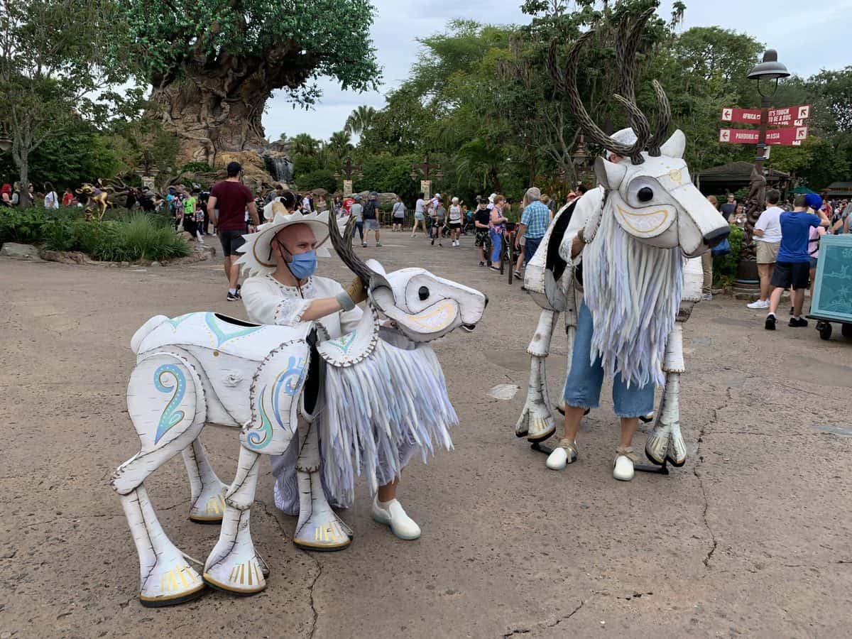 Whimsical white deer from the Merry Menagerie roam among guests at Disney’s Animal Kingdom, adding a magical seasonal flair to the park’s lively atmosphere with lush trees in the background.