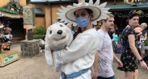 A whimsical performer in a festive white costume and hat delights guests by showcasing an adorable plush seal during Animal Kingdom’s Merry Menagerie celebration, while visitors enjoy the enchanting holiday atmosphere in the background.