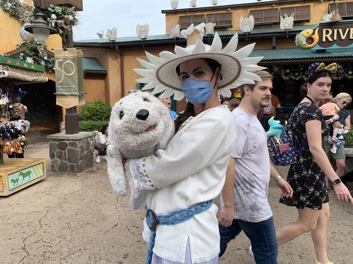 A whimsical performer in a festive white costume and hat delights guests by showcasing an adorable plush seal during Animal Kingdom’s Merry Menagerie celebration, while visitors enjoy the enchanting holiday atmosphere in the background.