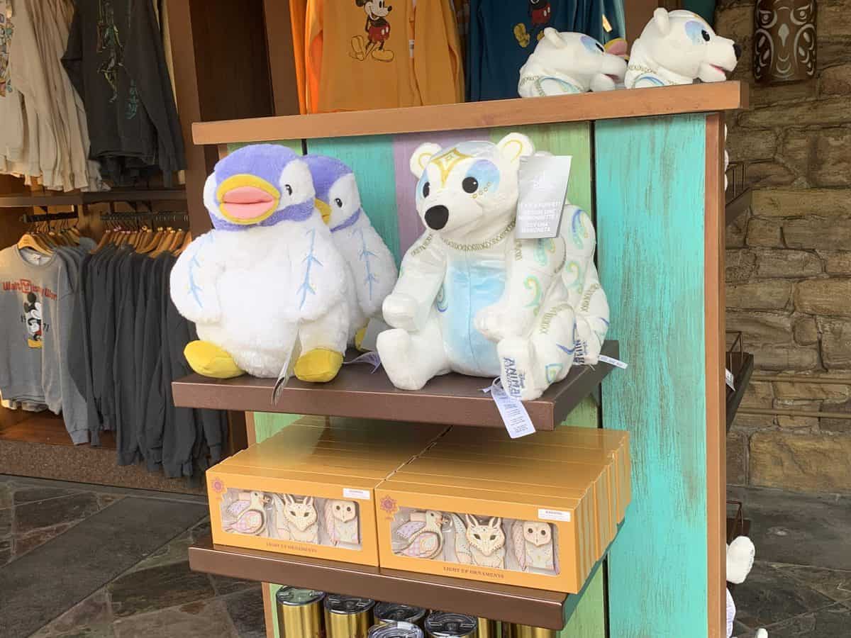 Merry Menagerie plush penguin and polar bear toys await guests on a display shelf at Disney’s Animal Kingdom, surrounded by festive gift boxes and souvenir shirts in the background.