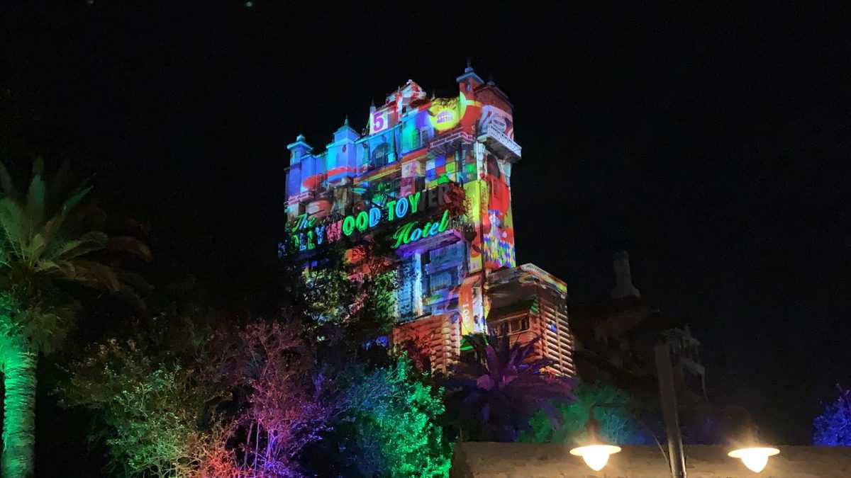 The Hollywood Tower Hotel on Sunset Boulevard at Disney's Hollywood Studios glows with vibrant, colorful lights during Sunset Seasons Greetings, as festive projections and sparkling displays transform the attraction while illuminated trees frame the scene in true holiday magic.