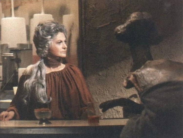 A gray-haired bartender stands behind the counter of a Star Wars-themed cantina, interacting with an alien animatronic featuring a long neck under moody, immersive lighting, evoking the atmosphere of the iconic Holiday Special scene.