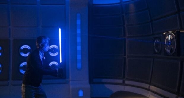 A Star Wars character wields a glowing blue lightsaber inside a dimly lit, futuristic attraction space featuring immersive blue accent lighting.