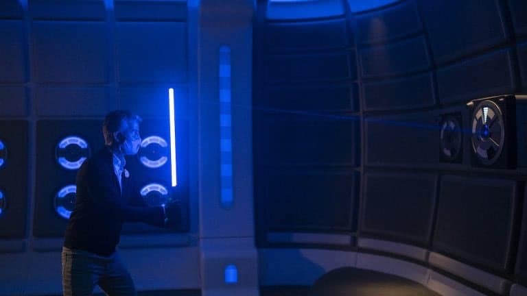 A Star Wars character wields a glowing blue lightsaber inside a dimly lit, futuristic attraction space featuring immersive blue accent lighting.