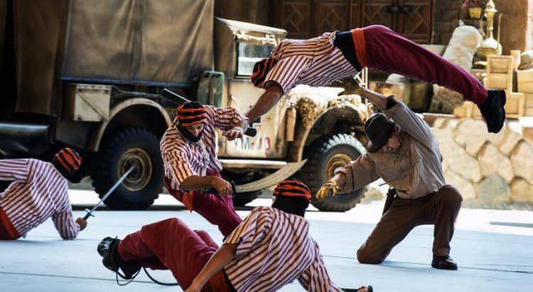 A man in a tan outfit dodges as four performers in red pants and striped shirts, some holding swords, enact an action scene; one performer is airborne above the man's head. A vintage truck and crates are in the background.