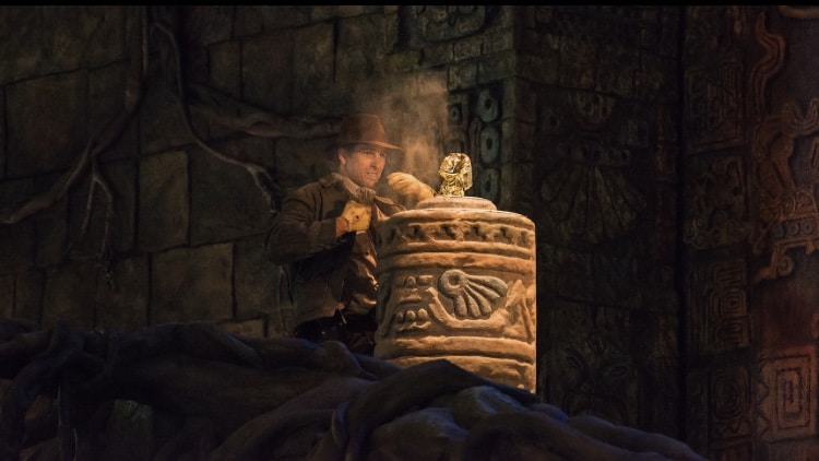 Indiana Jones, wearing his signature fedora, reaches for a golden idol inside a temple adorned with ancient carvings, capturing the excitement of the Indiana Jones Adventure attraction.
