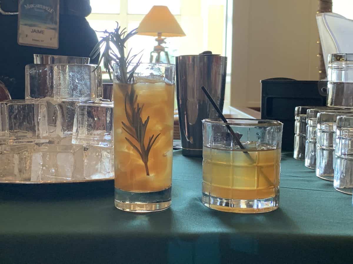 Holiday-inspired cocktails on a green tablecloth at Margaritaville Resort Orlando, one topped with rosemary, with sparkling ice cubes and stylish barware in the background—ideal for unwinding after a thrilling day at the resort’s attractions.