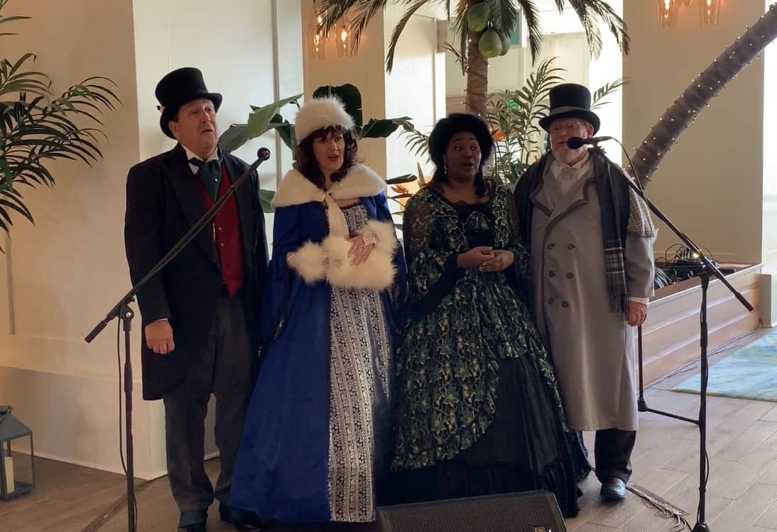 Victorian carolers perform festive holiday songs with microphones amid lush decor and greenery, entertaining guests at Margaritaville Resort Orlando.