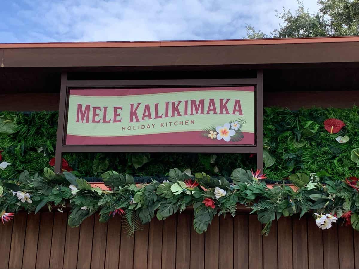 The Mele Kalikimaka Holiday Kitchen sign, adorned with tropical leaves and vibrant flowers, greets guests at EPCOT’s Festival of the Holidays.