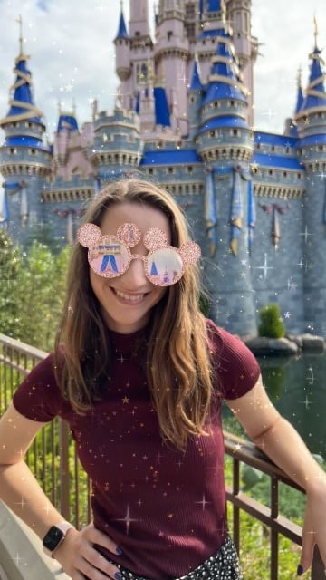 A Disney fan sporting Minnie Mouse ear sunglasses beams in front of Cinderella Castle, shimmering sparkles adding to the enchantment—an unforgettable memory made smoother with Disney Genie+ optimizing every magical moment at Walt Disney World.