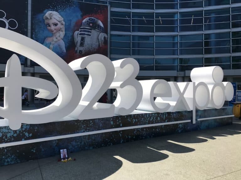 The bold white "D23 Expo" sign stands outside the convention center, with Elsa from Frozen and Baymax from Big Hero 6 featured on vibrant posters behind it, greeting Disney fans as they arrive for the excitement of Destination D23.