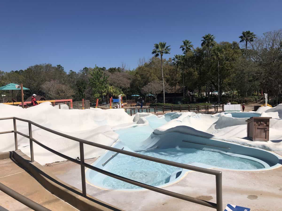 A closed pool with signature white, snowdrift-like edges inspired by Disney's Blizzard Beach is nestled among lush trees under a bright blue sky, capturing the iconic water park's wintry fun even when not in use.