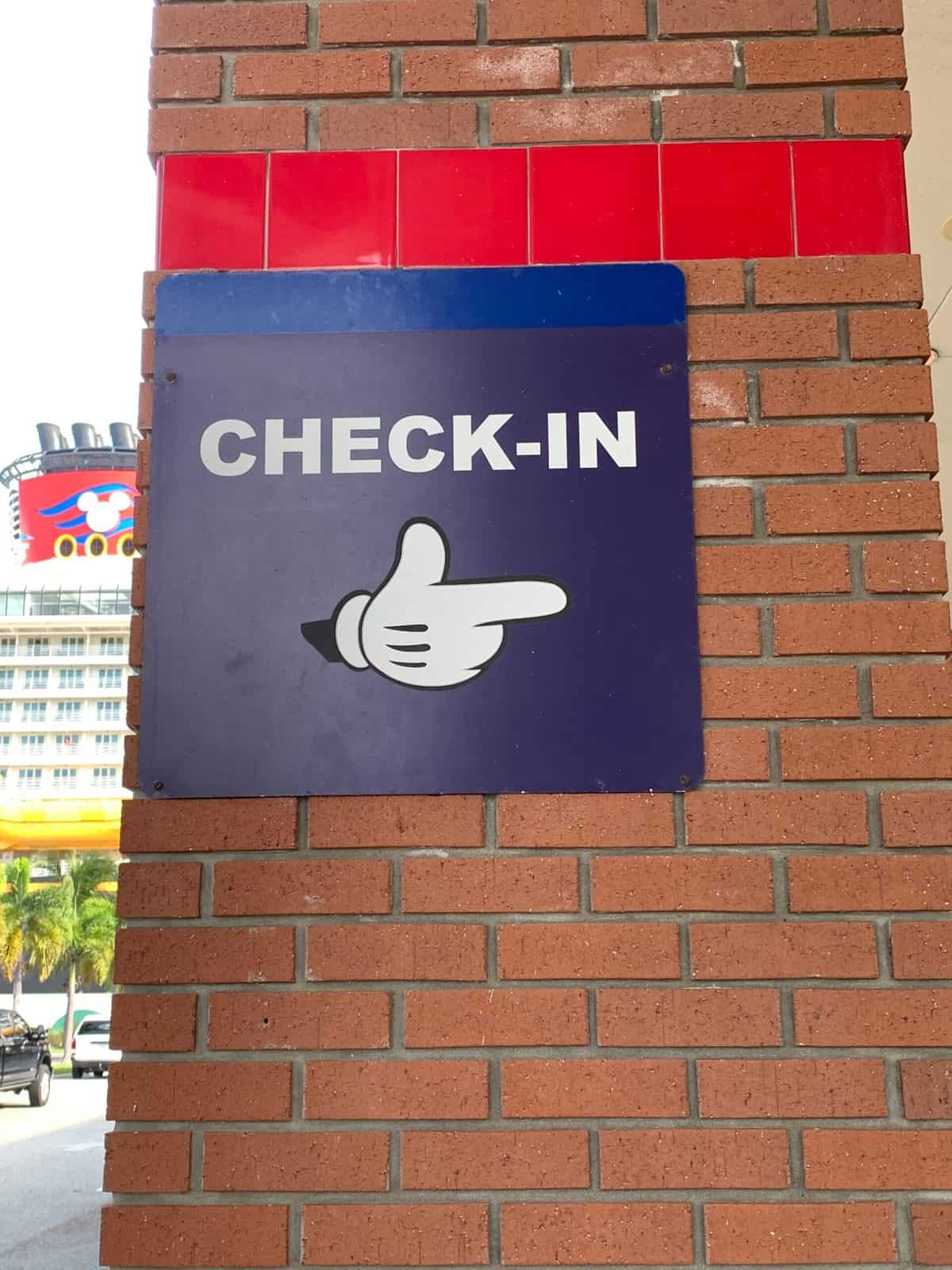 A playful purple "Check-In" sign with a cartoon hand pointing to the right, mounted on a brick wall—capturing the spirited atmosphere and magical anticipation of arriving at your Disney Cruise Line embarkation terminal.