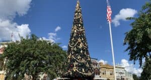 A towering Christmas tree shimmers with decorations in the center of Main Street, U.S.A., setting a magical holiday scene as park guests celebrate together. Nearby, the American flag flutters above, framed by sunshine—capturing that enchanting Disney Parks spirit where the wonders of the season come to life.