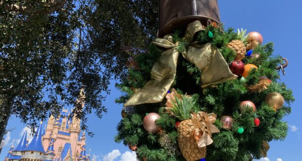 A festive wreath adorned with a gold bow and pineapples decorates a lamppost on Main Street, U.S.A. near Cinderella Castle at Magic Kingdom, set against a bright blue sky. The scene captures the enchanting holiday spirit of Disney Very Merriest After Hours, the exclusive sold-out event lighting up special nights at Walt Disney World.